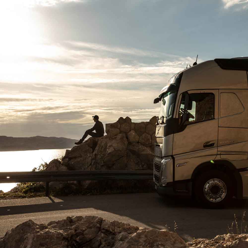 Kuljettaja istuu kalliolla katsomassa auringonlaskua merellä, Volvo FH -kuorma-auto pysäköitynä viereensä